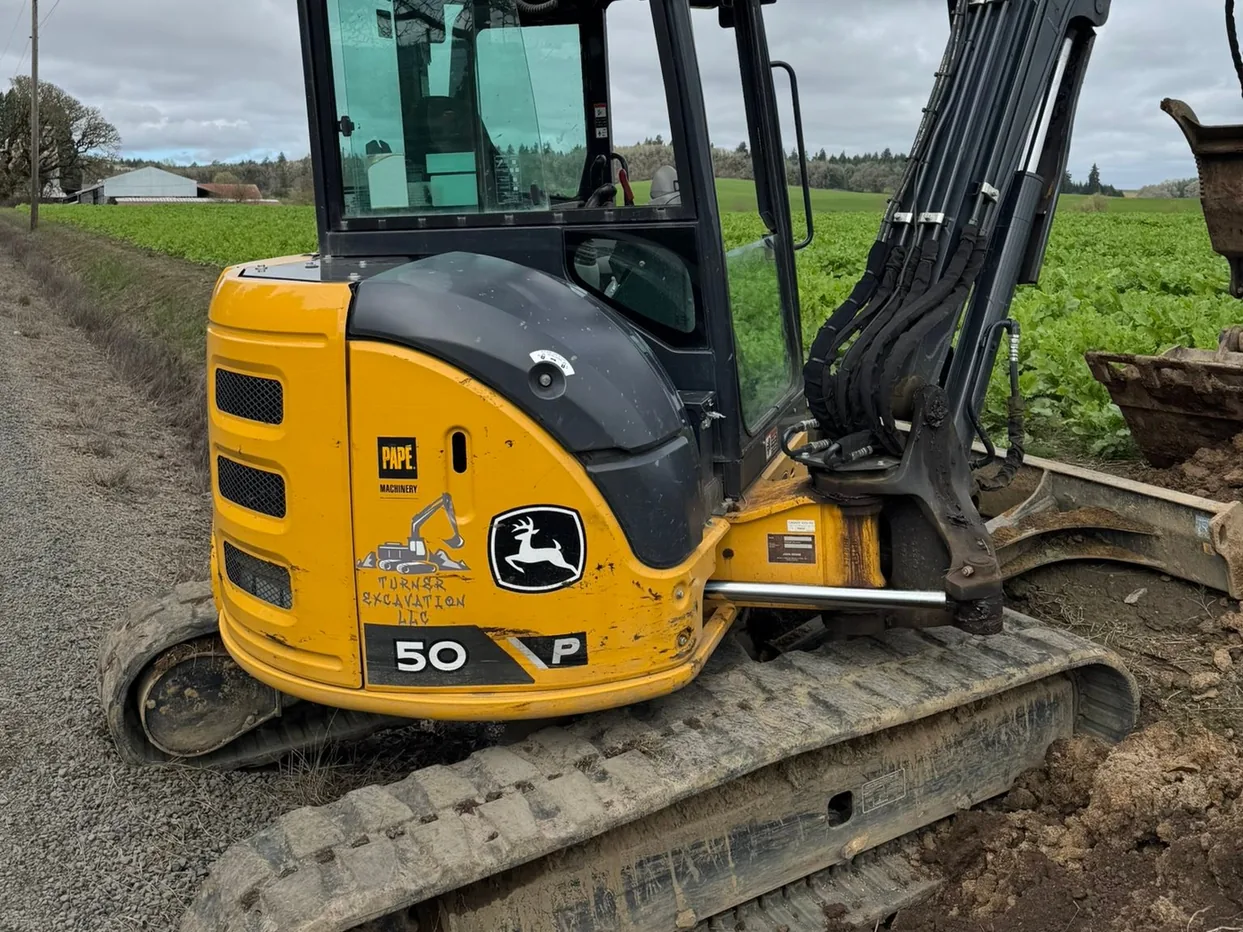 Compact excavator on a gravel edge beside a crop field, grading earthwork